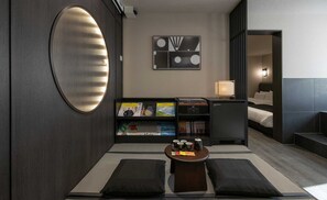 Une chambre d’hôtel moderne avec un grand miroir rond, une bibliothèque remplie de livres et de magazines, une petite table avec des tasses et un lit doté d’une tête de lit sombre.