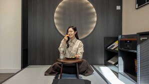 Una persona sentada a una mesa con un plato de comida, una bebida y un libro. La habitación tiene un diseño moderno con un elemento circular grande en la pared y una estantería.