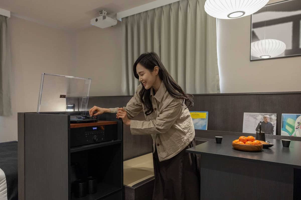 Una persona usando un dispositivo digital en una habitación de hotel con un escritorio, un cuenco de naranjas y un cuadro en la pared.