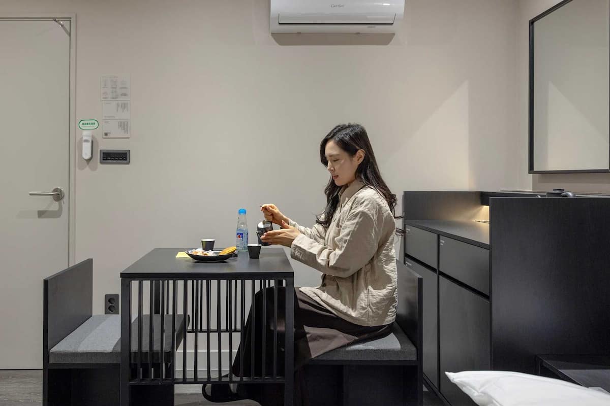 Habitación de hotel con una cama, un escritorio, una silla, una mesa con un plato de comida, una botella de agua y una persona usando un teléfono inteligente.