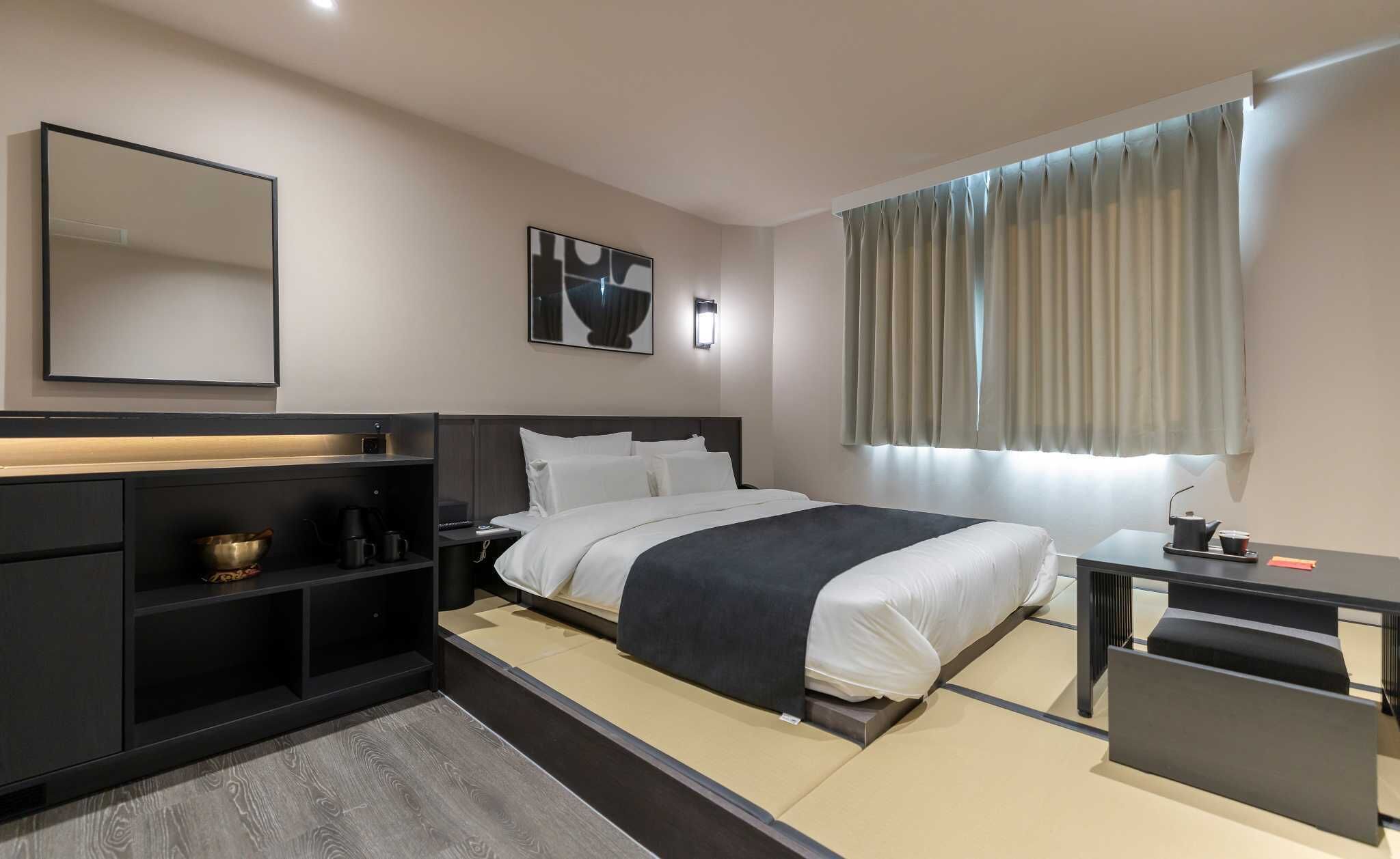 A modern hotel room with a bed, bedside table, desk, and a large window with curtains.