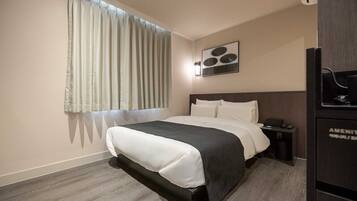 Habitación de hotel con cama, mesita de noche, escritorio, silla, ventana con cortinas y una obra de arte colgada en la pared.