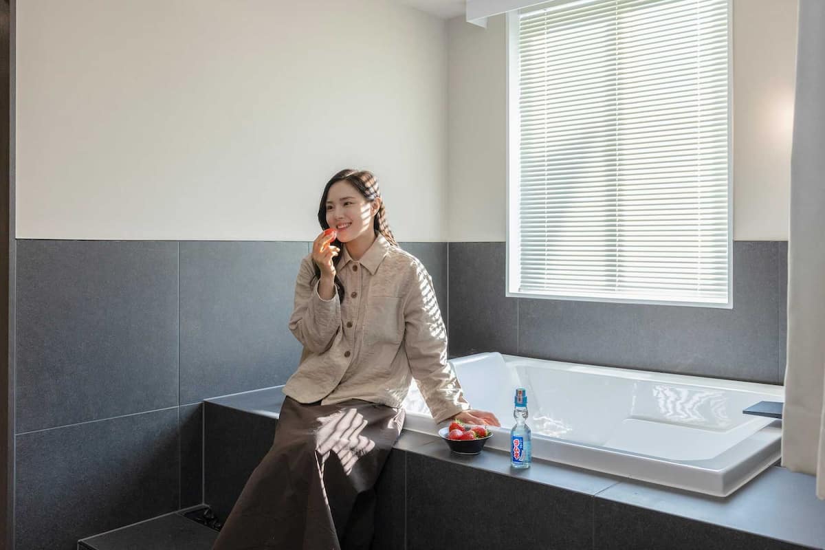 Una persona sentada en un banco en un baño moderno con una bañera grande, una ventana con persianas y una botella de agua al borde.