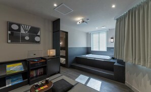 Quarto de hotel moderno com uma cama grande, uma escrivaninha com um laptop, uma estante de livros e uma janela ampla com cortinas.