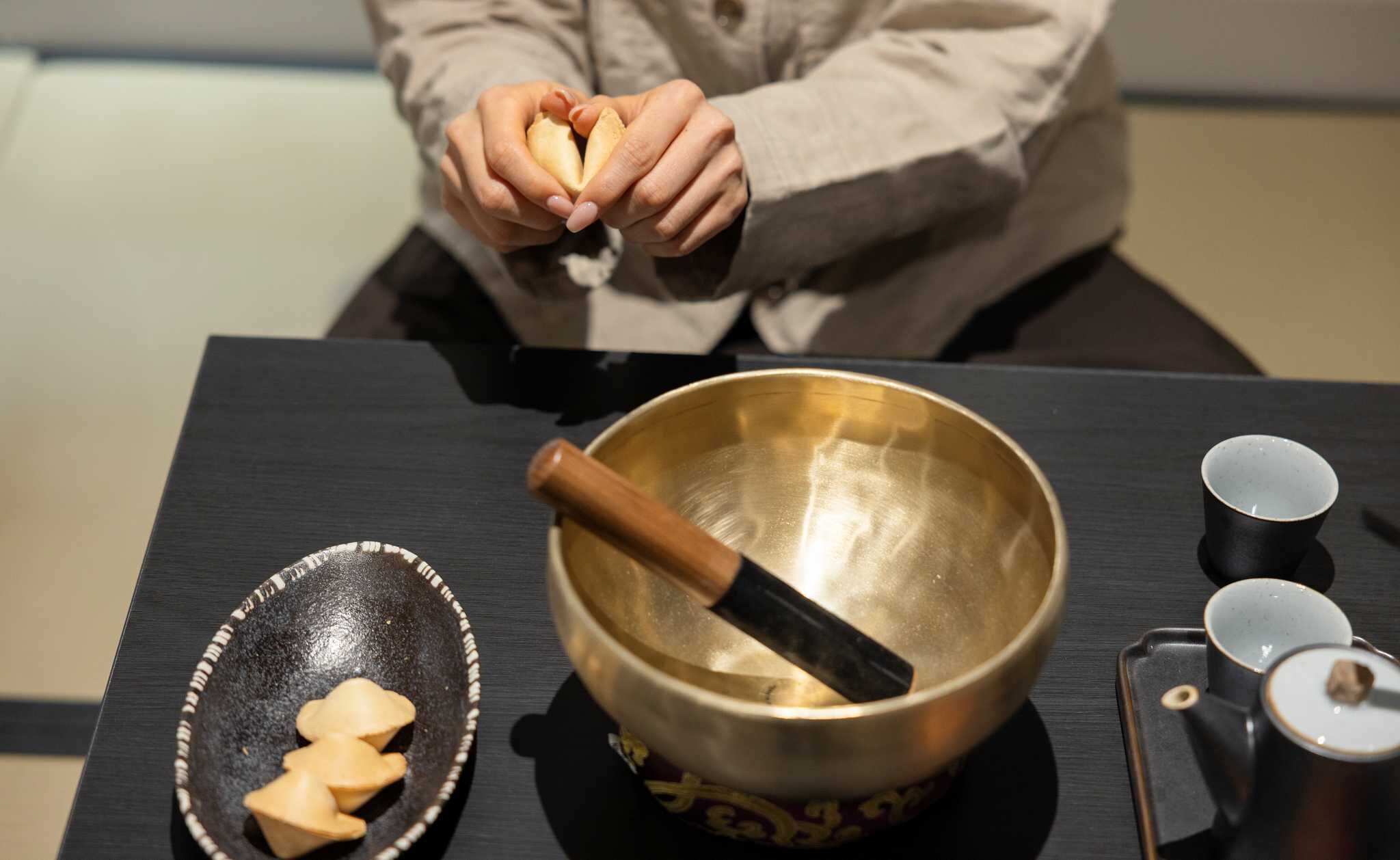 A person holding a small object, a golden bowl with a wooden mallet, and a black tray with three items on a table.