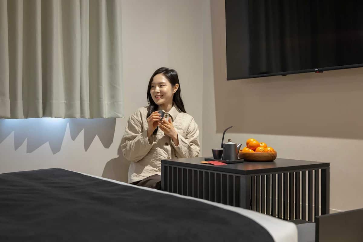 Una persona sentada en una cama en una habitación con un televisor montado en la pared, una mesita con un cuenco de fruta y un radiador.