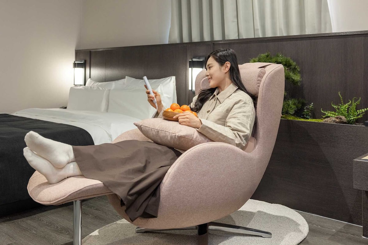 Una persona sentada en una silla moderna y acolchada con una computadora portátil, en una habitación que tiene una cama, una mesita de noche y una planta pequeña.