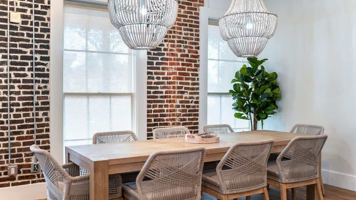 A dining area with a brick wall, large windows, wicker chairs, and a wooden table.