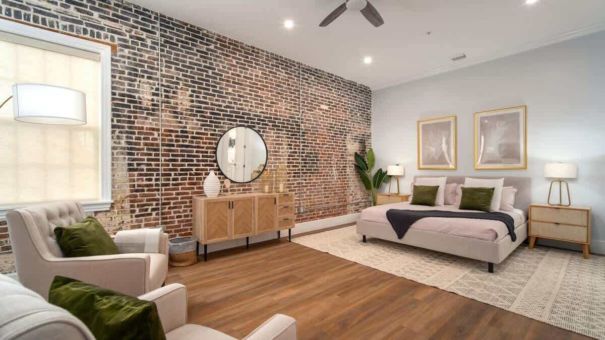 A modern bedroom with a brick wall, a round mirror, a bed with pillows, a nightstand, and a chair.