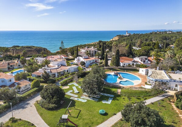 An aerial view of a coastal residential area with houses, a swimming pool, and a lighthouse in the distance.
