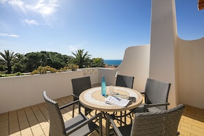 Ein runder Tisch mit Korbstühlen auf einem Balkon mit Blick auf eine malerische Meer- und Grünlandschaft.