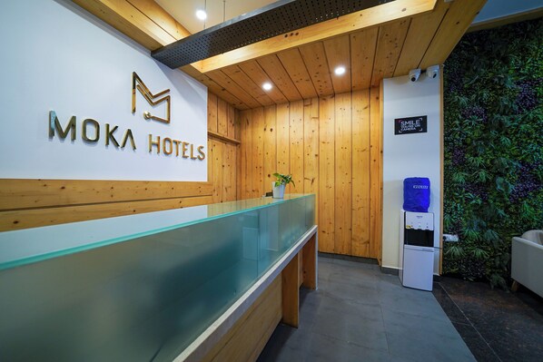 A reception area with a glass counter, wooden panel walls, a sign for Moka Hotels, a water dispenser, and a sign encouraging guests to smile. - Open by Olive - Moka Hotel (Bangalore)