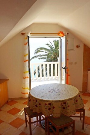Um quarto com uma mesa redonda coberta por uma toalha florida, duas cadeiras de madeira e vista para uma varanda com uma palmeira.