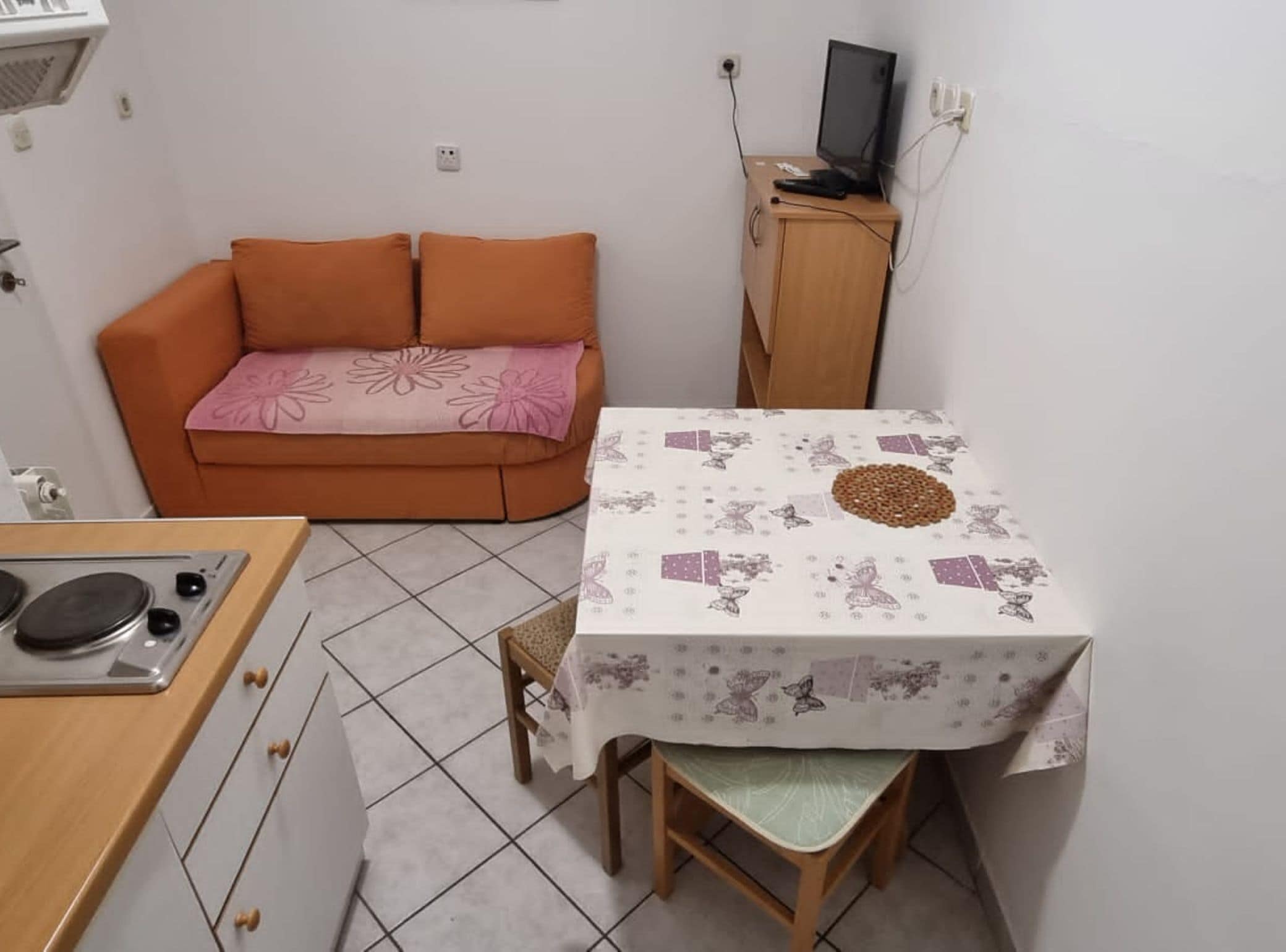 A compact living space with a kitchenette, a dining table with a floral tablecloth, an orange sofa with a pink cushion, and a wooden cabinet with a television.