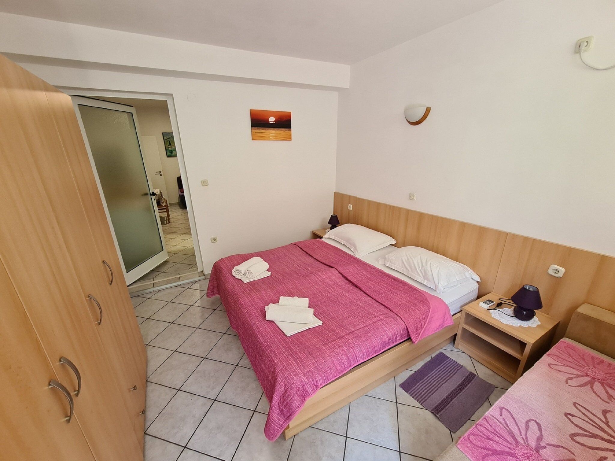 A hotel room with a bed, a nightstand, a lamp, a wall-mounted light, and a painting of a sunset.
