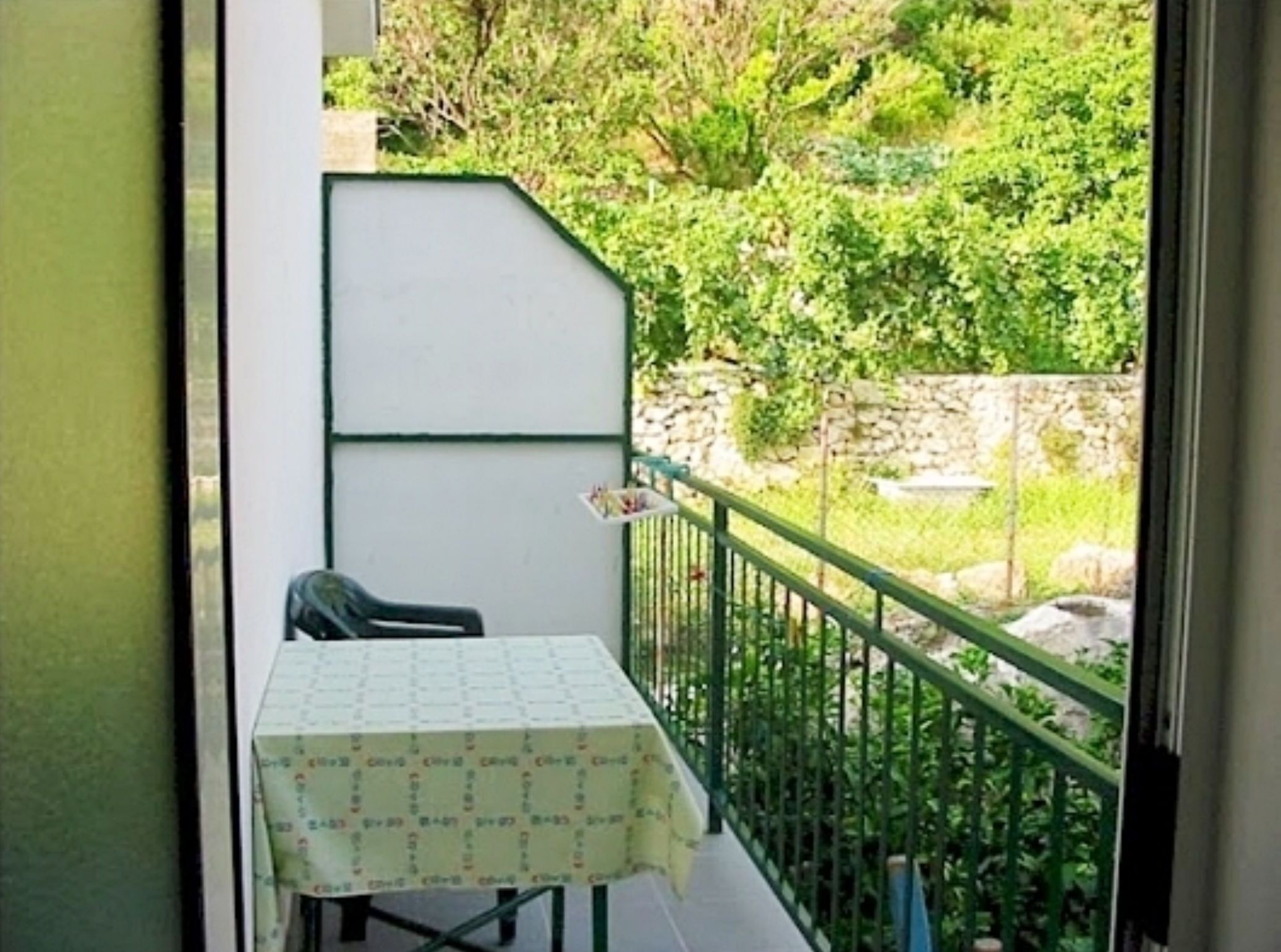 Un balcón con una mesa y una silla, barandilla verde y vistas a vegetación y un muro de piedra.