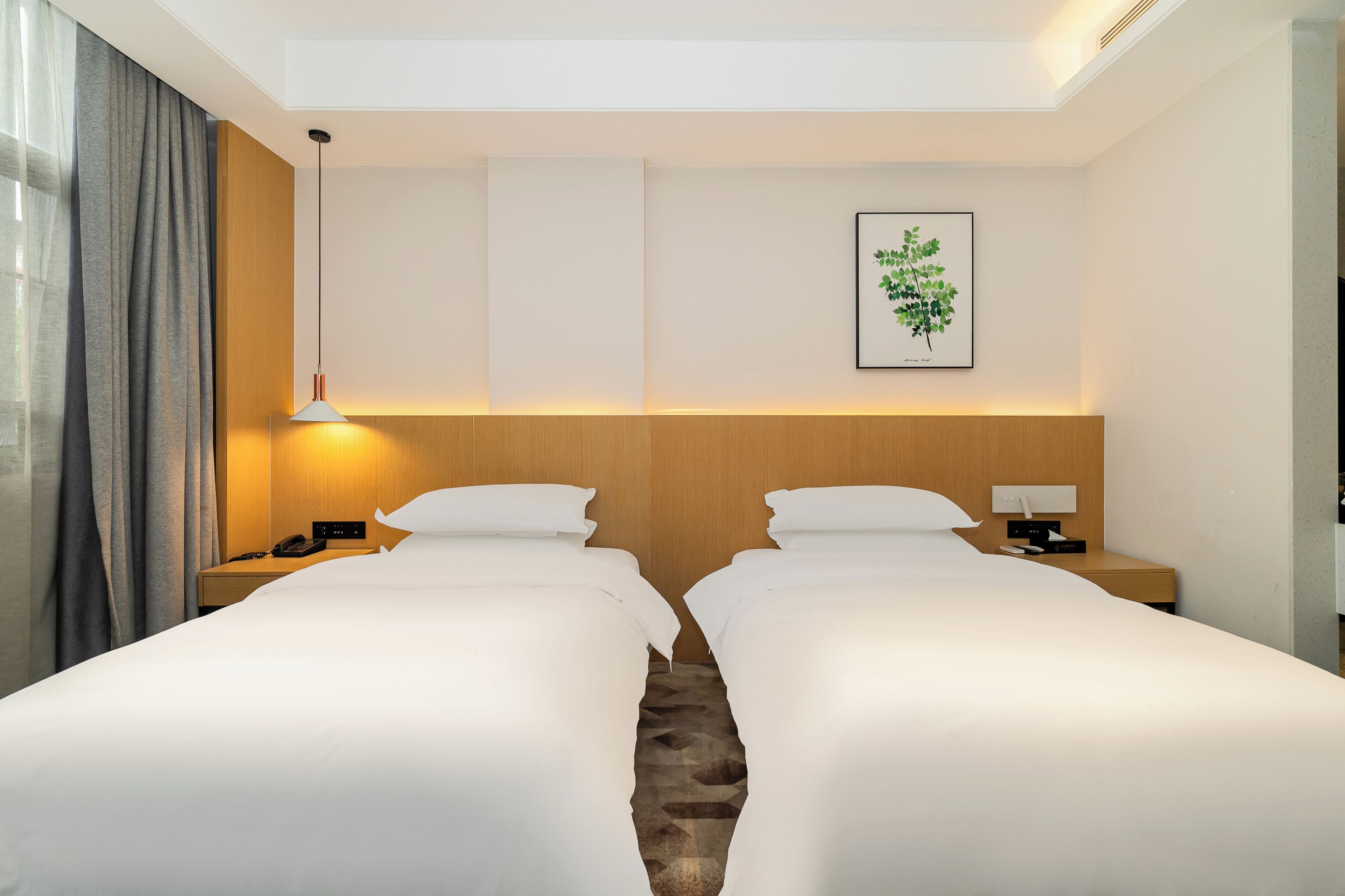 A hotel room with two beds, a bedside table with a lamp, a phone, and a wall-mounted light fixture.