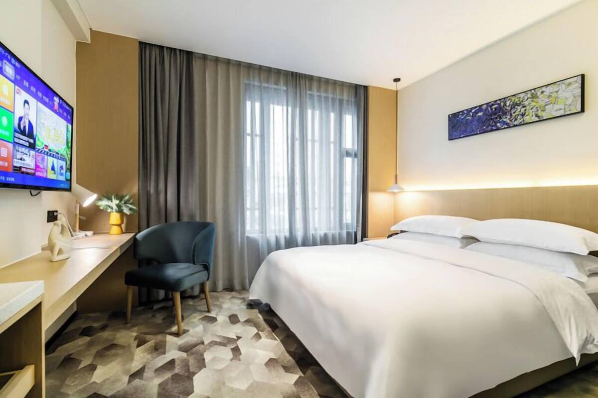Quarto de hotel com uma cama, escrivaninha com cadeira, televisão e janela com cortinas.