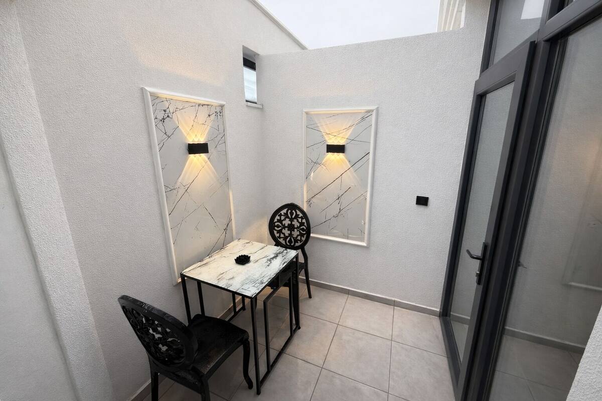 Un pequeño patio cerrado con mesa y sillas, dos luces fijadas a la pared y una puerta corrediza de vidrio.