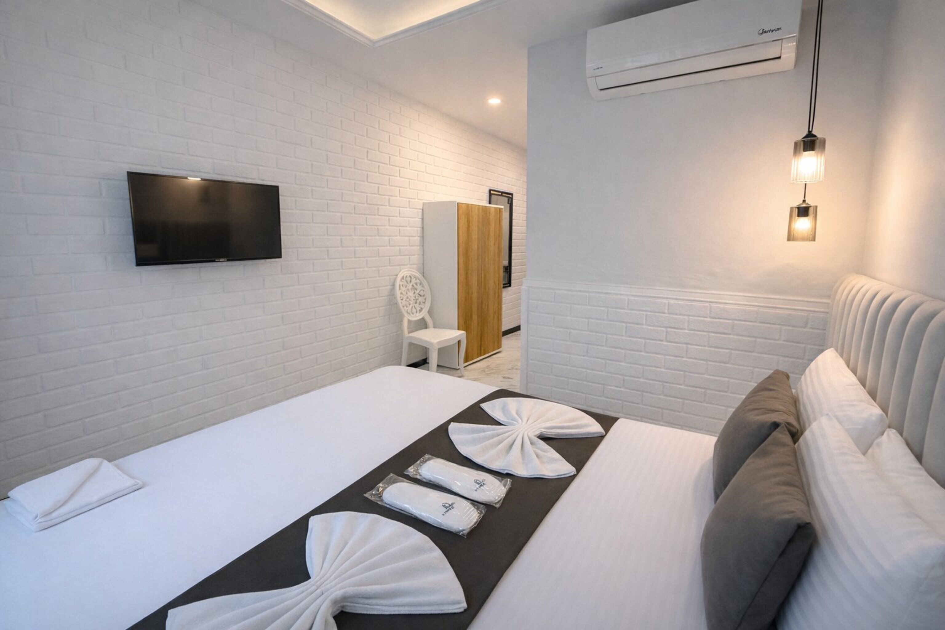 Une chambre d’hôtel dotée d’un téléviseur à écran plat fixé au mur, d’un lit avec du linge de lit blanc et des coussins décoratifs, d’une table de chevet avec une lampe et d’un mur en briques blanches.