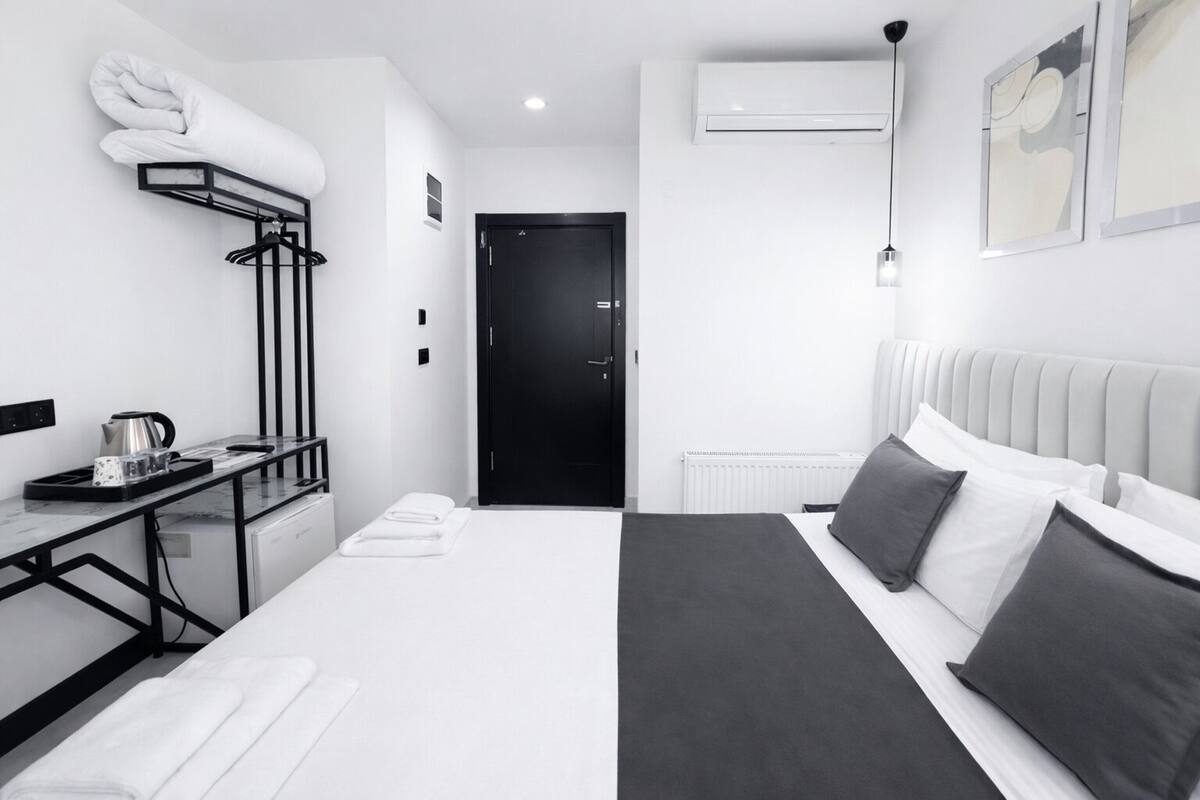 Una habitación de hotel moderna con una cama bien tendida, una mesita con una tetera, un radiador y una puerta negra.