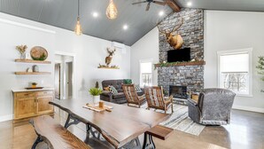 Un salon spacieux avec une grande table à manger en bois, des sièges confortables, une cheminée en pierre et un ventilateur de plafond.