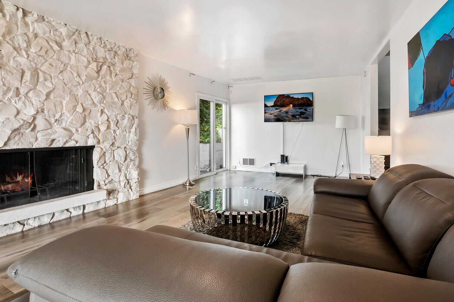 A modern living room with a stone wall, a fireplace, a glass coffee table, leather sofas, and a mounted TV.
