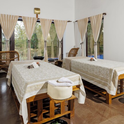 A room with two beds, wooden furniture, and large windows with curtains.
