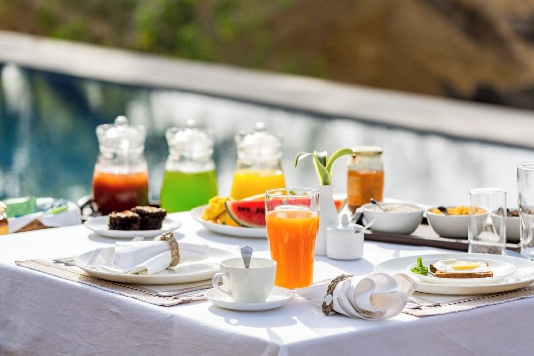 A breakfast setup by a pool with various drinks, fruits, and breakfast items on a table.