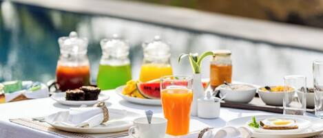 A breakfast setup by a pool with various drinks, fruits, and breakfast items on a table.