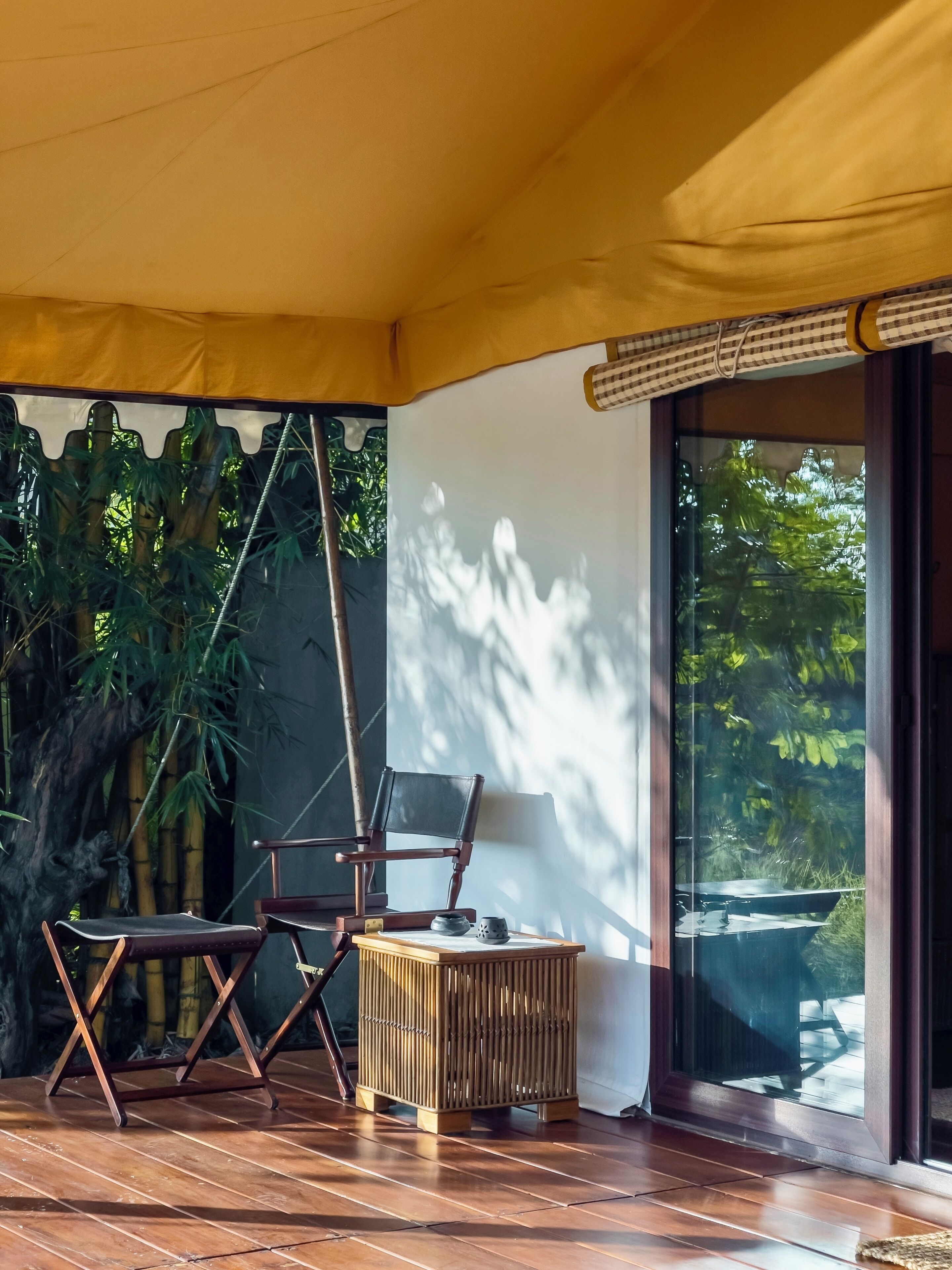 A wooden deck with two chairs and a small table, a bamboo stool, and a large window with a view of greenery.