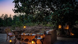 An outdoor dining area with wicker furniture and lanterns, surrounded by trees and vegetation. - Bagh Villas | Kanha (Baihar)
