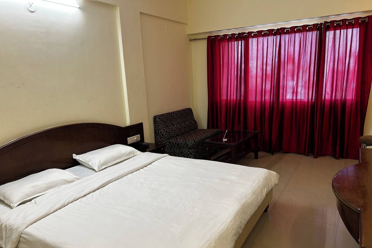 A hotel room with a bed, a chair, a small table, and a window with red curtains.