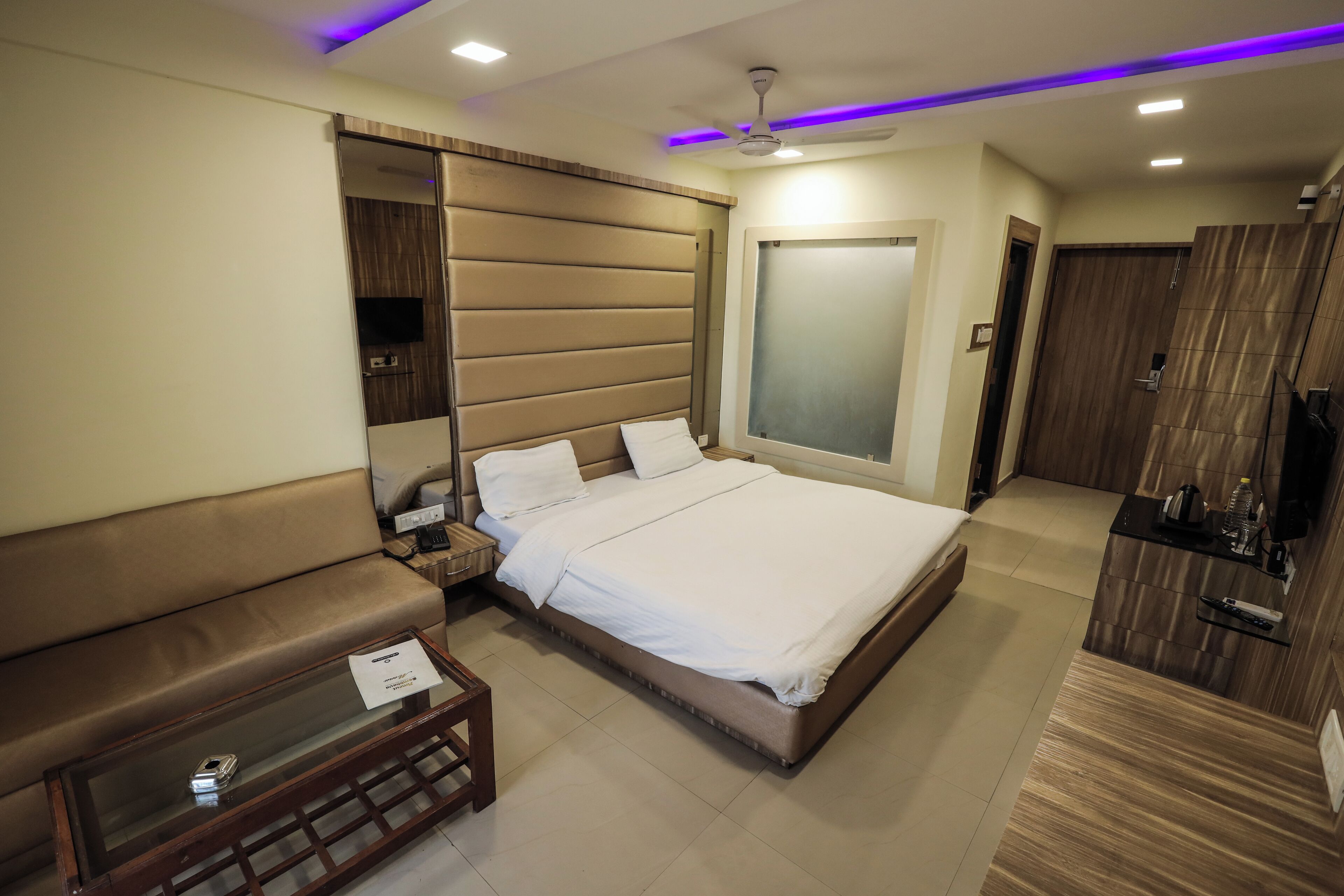 A modern hotel room with a large bed, a sofa, a glass coffee table, and a TV on a wooden cabinet.