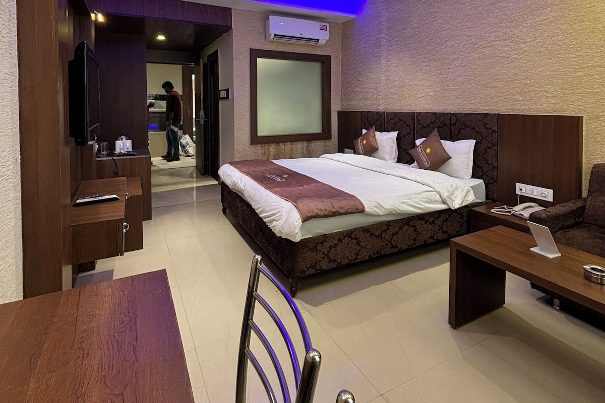 A hotel room with a large bed, a desk, a chair, a TV, and an air conditioning unit.