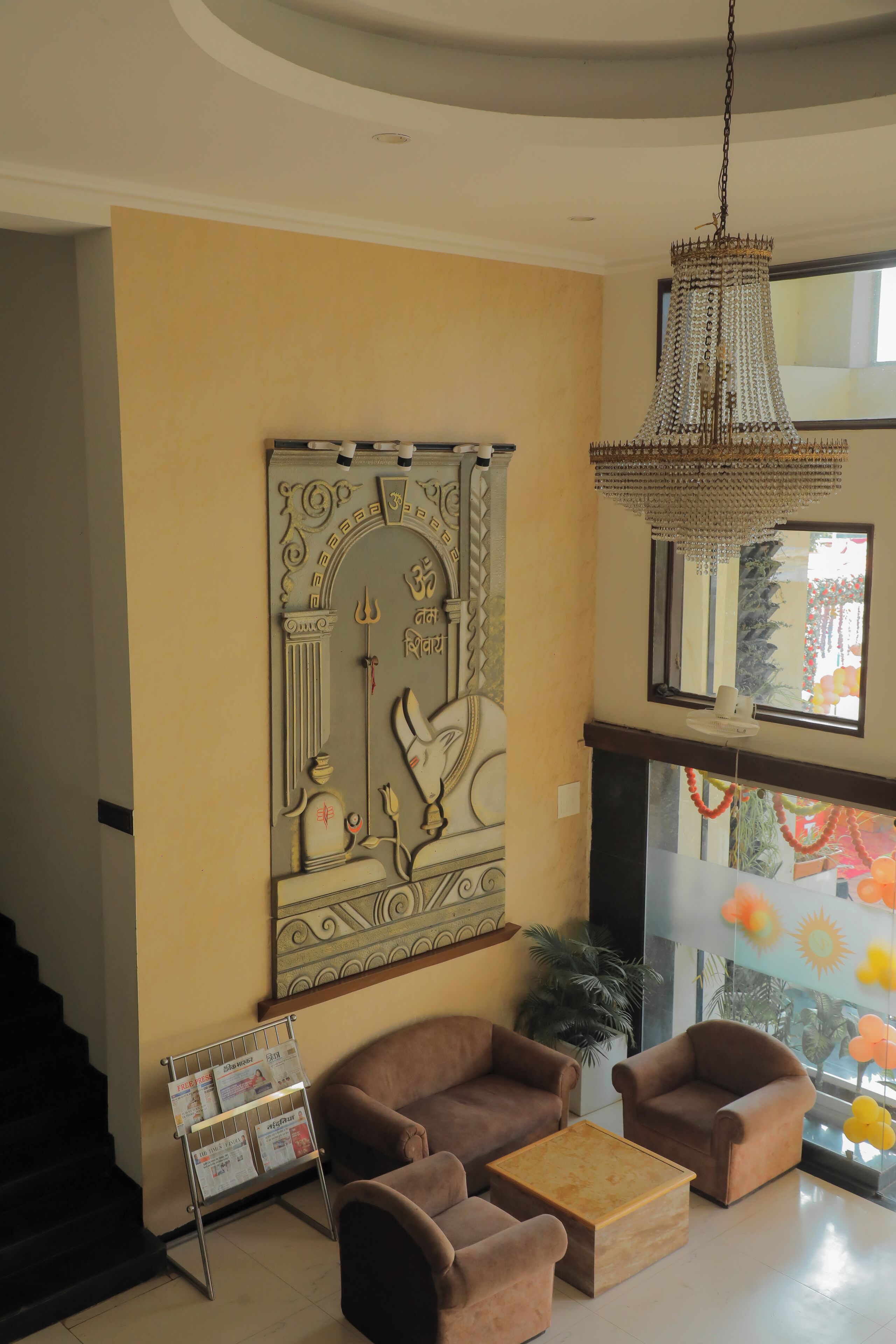 A lobby with a reception desk, two brown armchairs, a wooden coffee table, a chandelier, and a decorative wall piece with a deity figure.