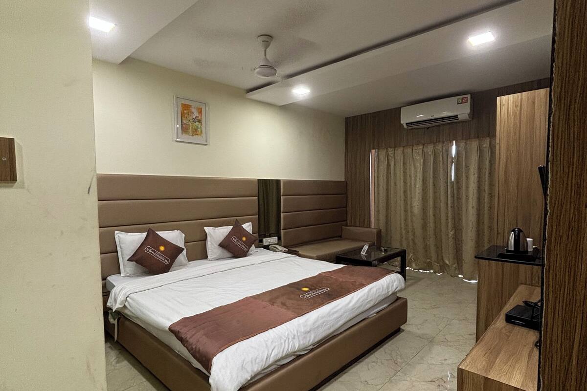 A hotel room with a large bed, a wooden headboard, a bedside table, a desk with a chair, a mirror, an air conditioning unit, and a window with curtains.