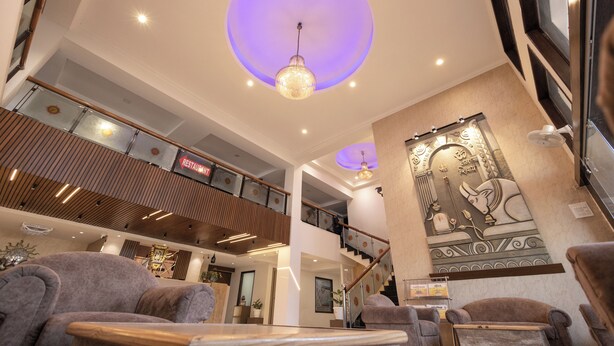 A spacious lobby with a high ceiling, a central chandelier, and a staircase leading to an upper level. - Hotel Vikramaditya (Ujjain)