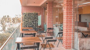 An outdoor seating area with wooden tables and chairs, a brick wall, and a glass partition. - Hotel Vikramaditya (Ujjain)