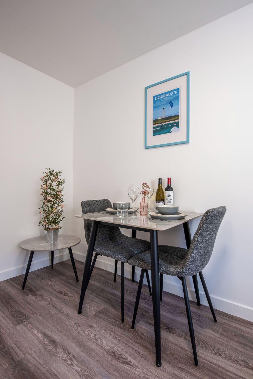 Comedor con mesa dispuesta para cuatro, con un cuadro enmarcado, botellas de vino y copas.