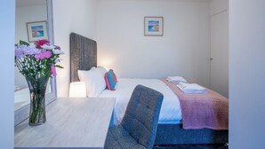 A hotel room with a bed, a desk, a chair, a mirror, and a vase of flowers. - Harbour Nights Deluxe 7E by Interhome (Lossiemouth)
