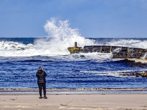 Una persona está de pie en una playa arenosa, mirando olas oceánicas agitadas chocando contra una estructura de concreto.