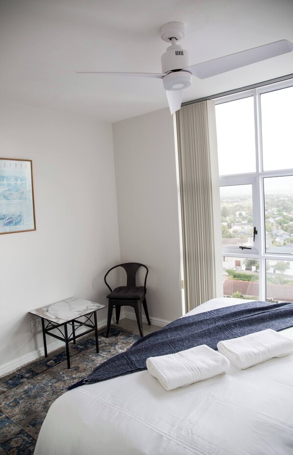 A hotel room with a bed, a desk, a chair, a ceiling fan, and a large window with a city view.