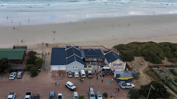 Vista aérea de un edificio frente al mar, con estacionamiento, una tienda de surf y gente caminando por la playa.