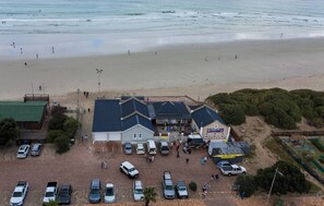 Et luftfoto af en bygning ved stranden med en parkeringsplads, en surfbutik og mennesker, der går på stranden.