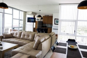 Ein modernes Wohnzimmer mit einem großzügigen L-förmigen Sofa, einem Esstisch mit Platzsets und einer Küchenzeile mit Holdekor und einem Fernseher.