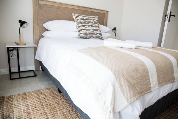 A neatly made bed with white and beige bedding, a patterned pillow, and a bedside lamp on a small table beside the bed.