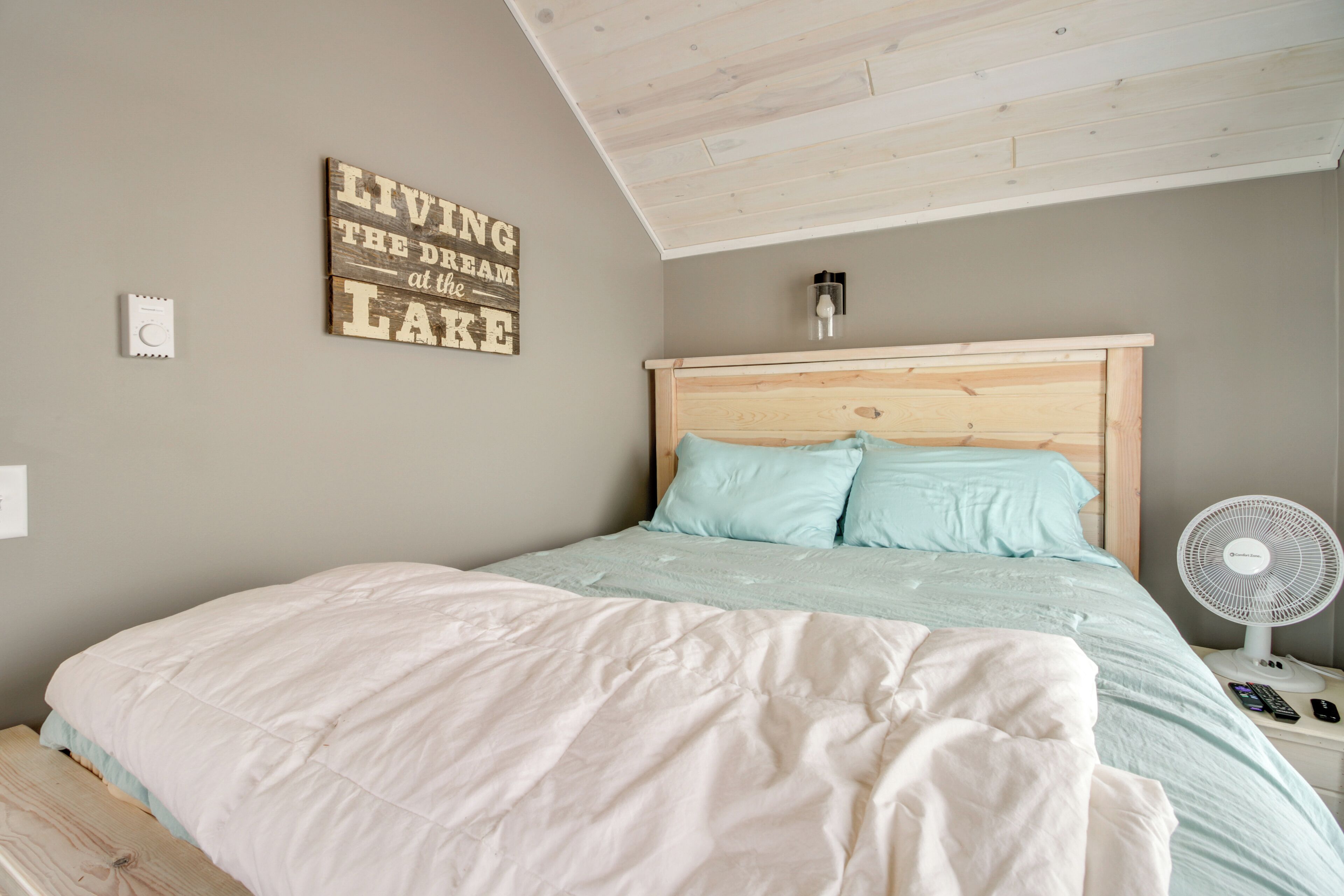 Une chambre à coucher avec une tête de lit en bois, un lit recouvert d’une couverture blanche et bleu clair, une petite table de chevet avec un ventilateur et un luminaire fixé au mur.