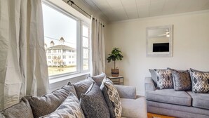 Ein Wohnzimmer mit grauem Sofa, gemusterten Kissen, einer Zimmerpflanze und Blick auf ein Gebäude.