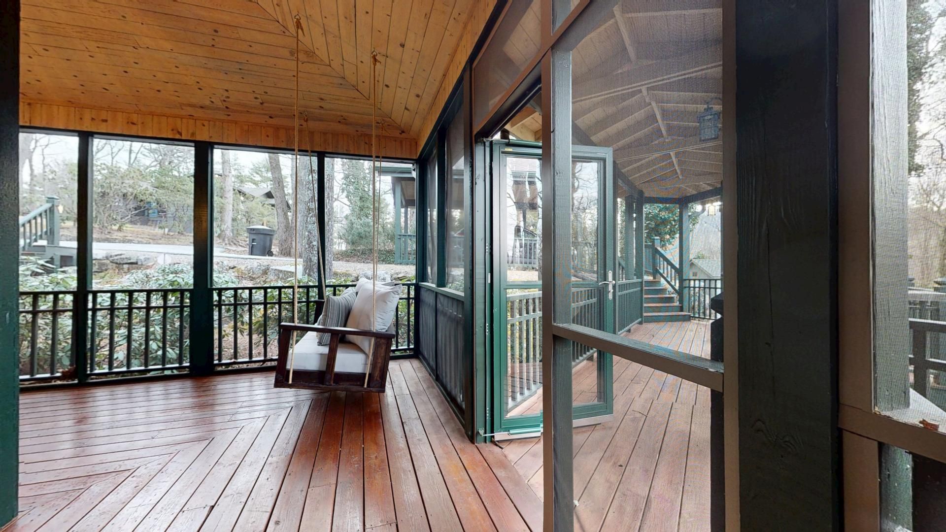 A covered porch with wooden flooring, a cushioned chair, and large windows offering views of the outdoors.